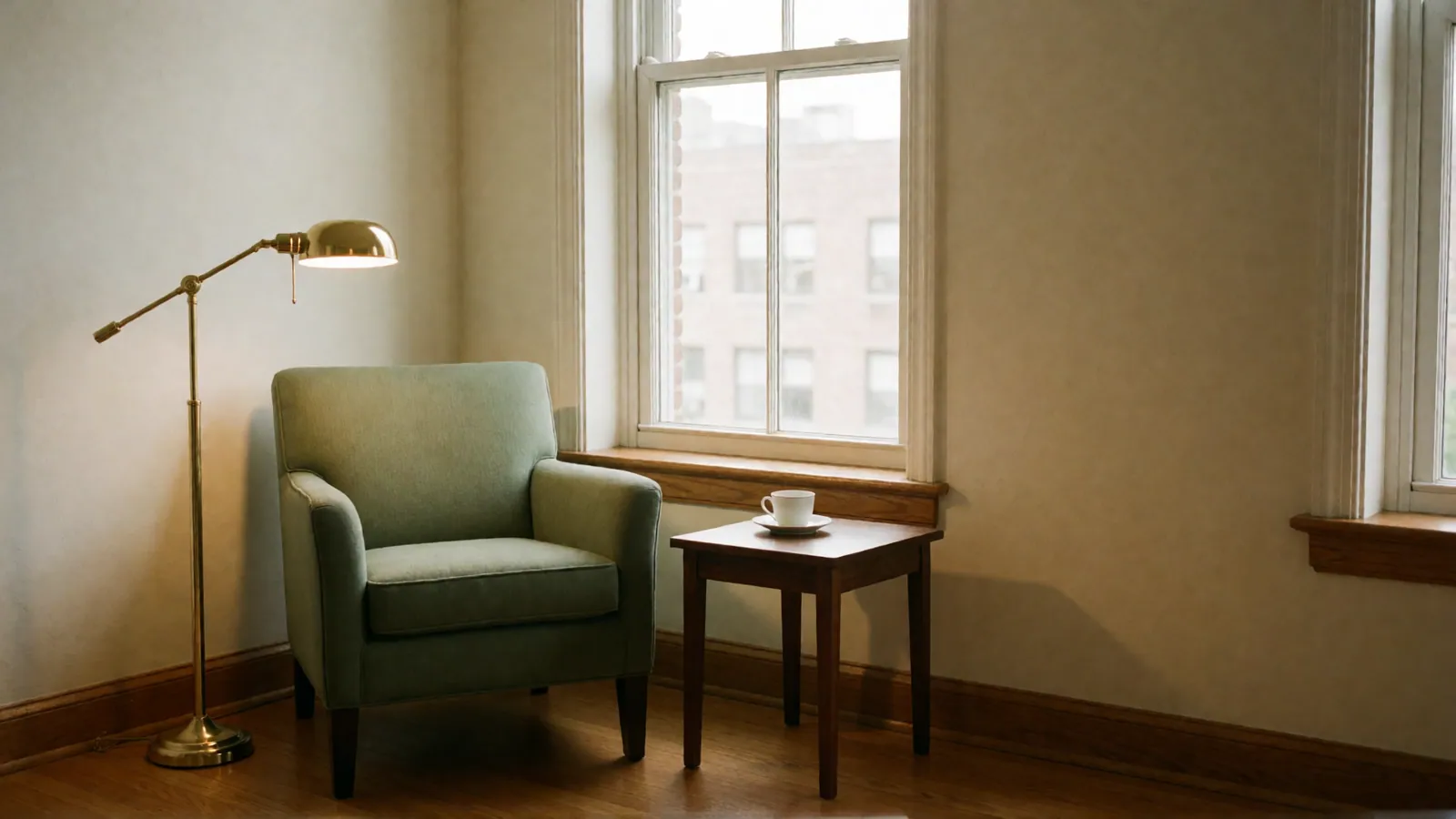 A brass floor lamp lit beside an empty green armchair and a small side table with a cup and saucer, by a double-hung window.