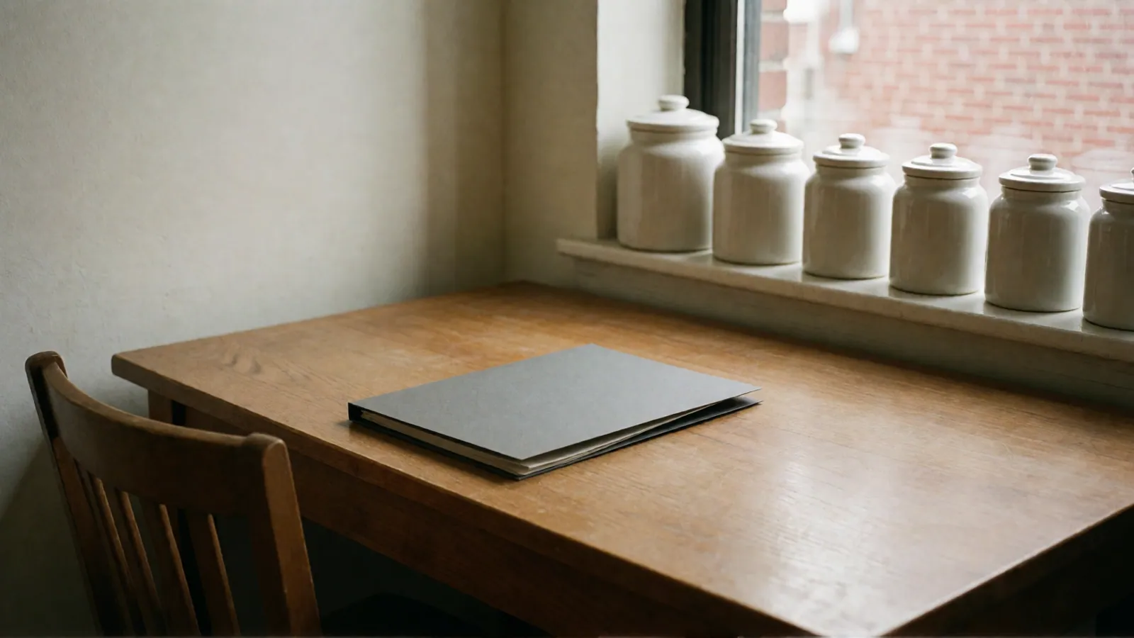 A plain wooden worktable with a closed gray folder and a wooden chair, a row of lidded white ceramic jars along the windowsill and red brick through the window.
